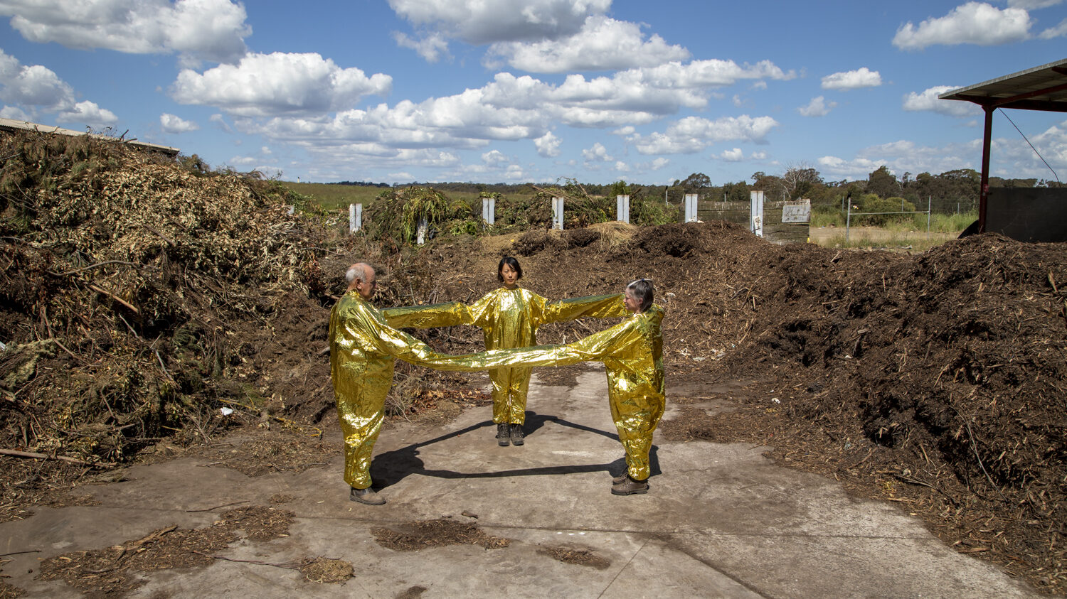 Three people standing outside in gold boiler suits holding hands in a triangle shape.