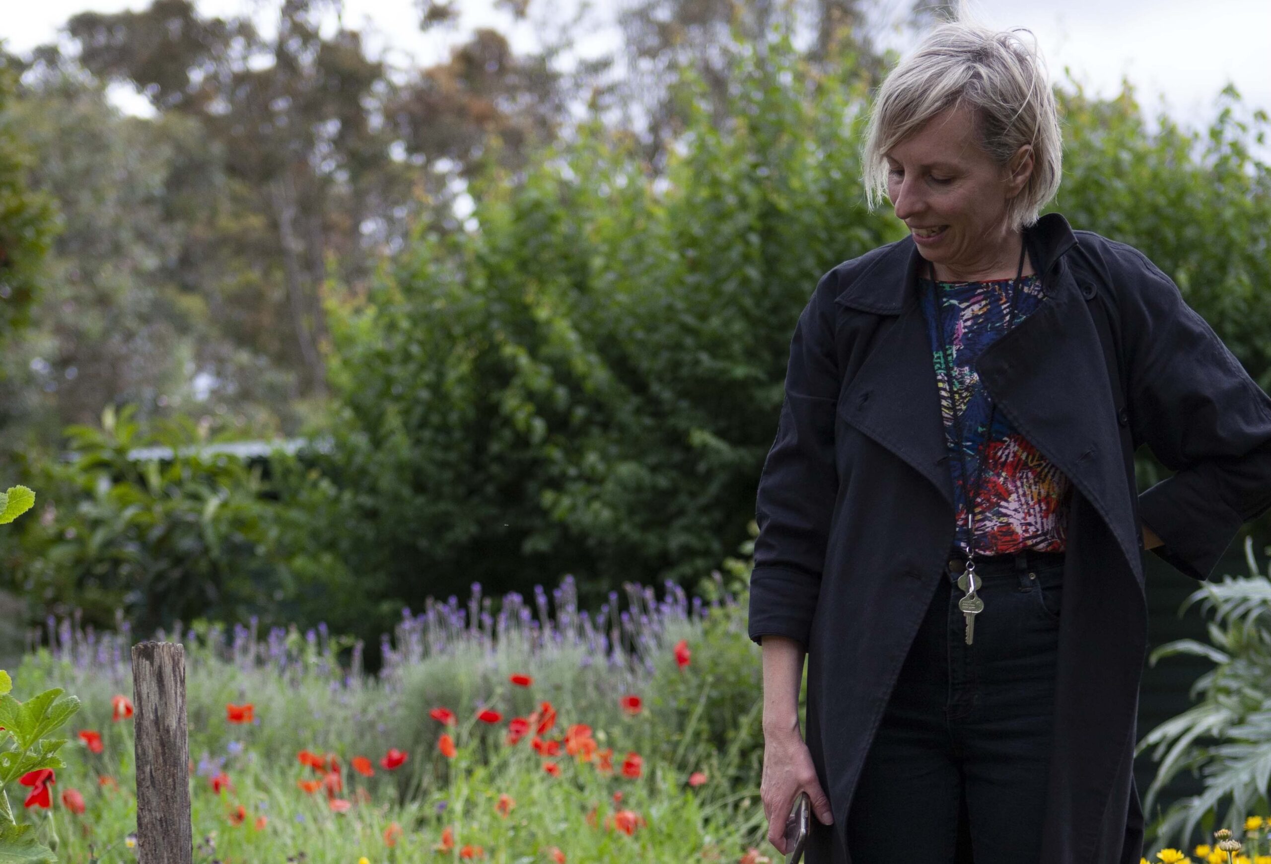 An image of RAV's Communications and Development Coordinator Claire Miovich. Clare has short blonde hair and wears a long black coat. She is standing in a lush garden with poppies and lavender in the background. In the distance there are some out of focus eucalyptus trees.