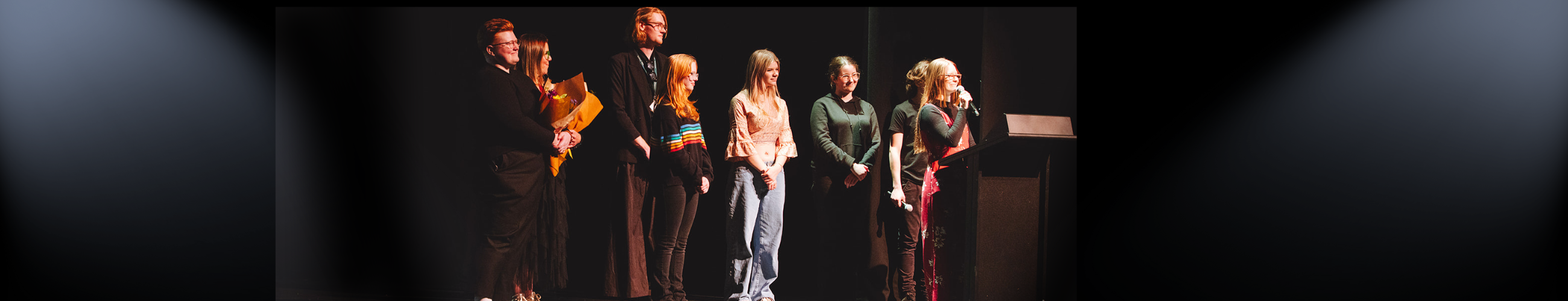 A group of young people on stage, one talking at a lecturn